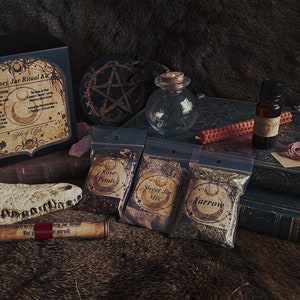 Könnte beinhalten: Ein Honey Jar Ritual Kit mit verschiedenen Komponenten. Enthält eine schwarze Box mit Text, kleine Glasflaschen, Beutel mit Kräutern mit der Aufschrift "Rose Petals", "Sweet Jar Mix" und "Yarrow", eine Schriftrolle und ein Pentakel. Das Kit ist auf einer strukturierten Oberfläche angeordnet.
