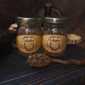 Może przedstawiać: Dwa szklane słoiki ze srebrnymi pokrywkami, z napisem "Lucifer Loose Incense Blend", stoją na ciemnoniebieskiej powierzchni. Drewniana łyżka zawiera luźne kadzidło. Etykiety są beżowe z czarnym tekstem.