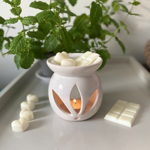May include: White ceramic wax warmer with a candle lit inside. The warmer has a decorative leaf design and is filled with white wax melts in the shape of hearts. There are additional wax melts on the surface next to the warmer.