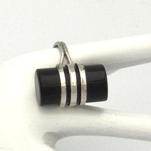 May include: A silver ring with a black, cylindrical centerpiece. The black cylinder is accented with three silver bands. The ring is displayed on a white surface, showcasing its design and details.