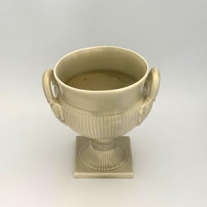 May include: A cream-colored urn with two handles and a square base. The urn has a fluted body and decorative relief details near the handles. The interior of the urn appears to contain liquid. This decorative piece is suitable for home decor.