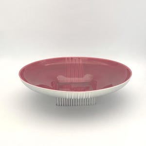May include: A decorative serving bowl with a wide, shallow, oval shape. The interior is a deep red, contrasting with the white rim and base. The base has a vertical ribbed design. A unique piece for serving or display.
