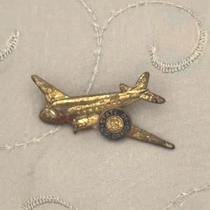 May include: A vintage gold-toned airplane pin with a small circular emblem. The pin features a detailed design of an airplane, with wings and a tail. The emblem has the words "STATE OF COLORADO" around the edge.