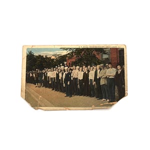 May include: Vintage postcard featuring a black and white photograph of a group of people standing in a line. The postcard has a faded appearance, with a building and trees in the background. The text on the postcard is blurry.