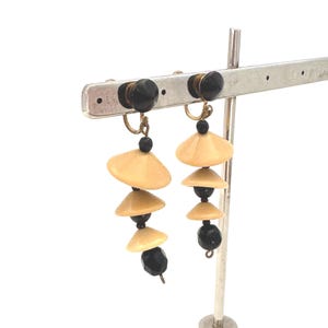 May include: A pair of vintage clip-on earrings. Each earring features a series of cream-colored, disc-shaped elements and black beads, suspended from a black and gold clip. The earrings are displayed on a silver earring stand.