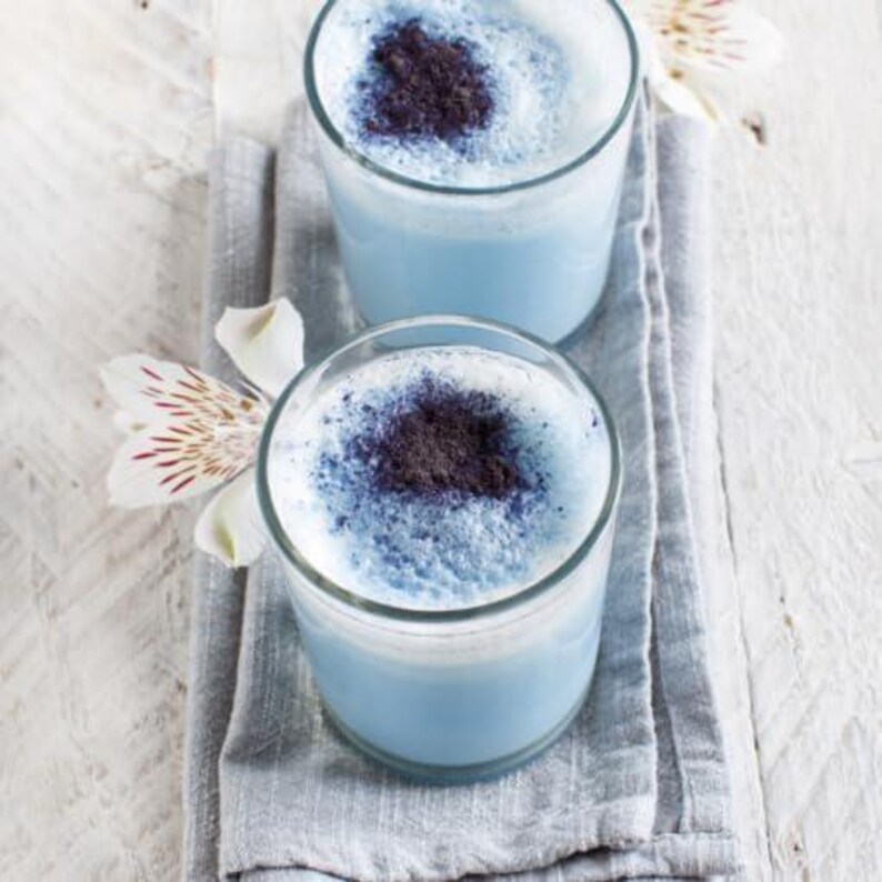 May include: Two clear glasses filled with a light blue beverage, topped with a dark powder. The drinks sit on a gray napkin, with white and red-streaked flowers in the background. The glasses are on a white wooden surface.