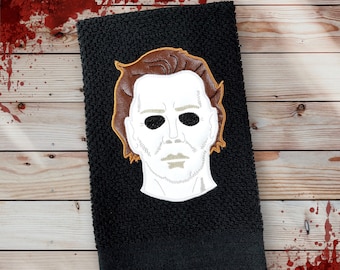May include: Black kitchen towel with an embroidered white face mask with brown hair. The mask has a stitched-on white mouth and eyes.