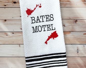 May include: White kitchen towel with black and white stripes at the bottom. The towel has red blood splatter and the text "Bates Motel" embroidered in black.