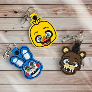 May include: Three embroidered keychains featuring characters from the Five Nights at Freddy's video game series. The keychains are yellow, blue, and brown, and each features a different character's face.