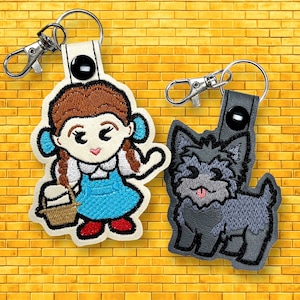 May include: Two embroidered keychains featuring characters from The Wizard of Oz. One depicts Dorothy in a blue dress with a basket, the other a gray dog. Both have silver-tone metal clasps and are set against a yellow brick background.