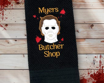 May include: Black hand towel with the embroidered text "Myers Butcher Shop" in gold. The towel features an embroidered image of a white-masked face with brown hair and red accents.