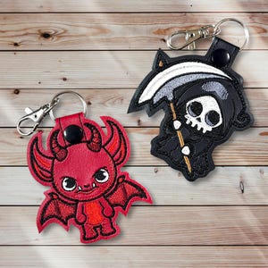 May include: Two embroidered keychains featuring cartoon characters. One is a red devil with horns and wings, the other is a black Grim Reaper with a scythe. Both have silver key rings and black leather accents.