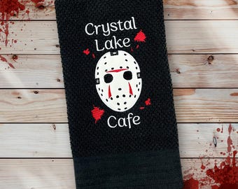May include: Black kitchen towel with the words "Crystal Lake Cafe" and a white hockey mask design with red accents. The towel is made of a textured fabric.