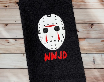 May include: Black hand towel with an embroidered white hockey mask with red accents. The text "WWJD" is embroidered in red below the mask. The towel is displayed on a wooden surface.
