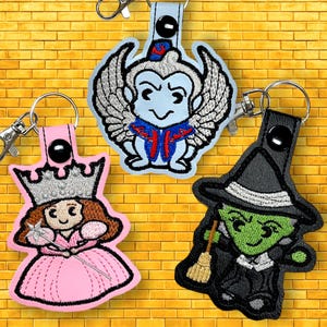May include: Three Wizard of Oz character keychains. The Wicked Witch of the West is green with a black dress and hat, holding a broom. The winged monkey is blue with a red and blue uniform. The Good Witch of the North is pink with a silver crown and a wand.