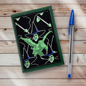 May include: A green leather notebook with a black and green fabric cover featuring a repeating pattern of the Wicked Witch of the West from the Wizard of Oz. The witch is wearing a black hat and green dress and is flying on a green monkey.