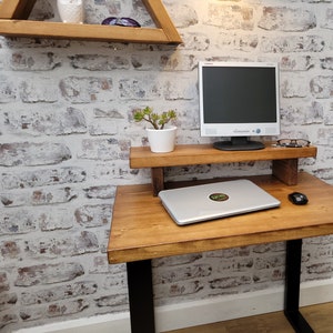 May include: A wooden desk with a black metal frame and a wooden monitor stand. A laptop computer, a mouse, and a potted plant are on the desk. The desk is in front of a white brick wall.