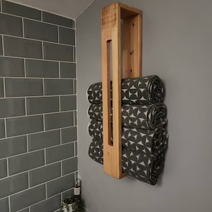 May include: A vertical wall-mounted wooden towel rack with a light brown natural wood finish. The rack holds four rolled towels featuring a dark gray and white geometric pattern consisting of repeating circular and triangular shapes.