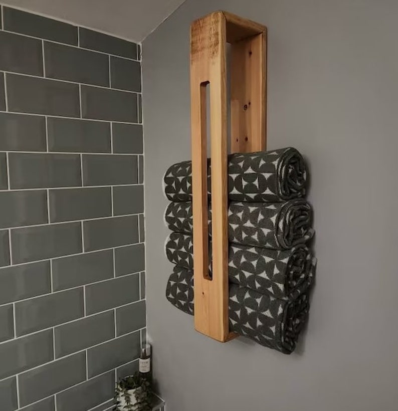 May include: A vertical wall-mounted wooden towel rack with a light brown natural wood finish. The rack holds four rolled towels featuring a dark gray and white geometric pattern consisting of repeating circular and triangular shapes.