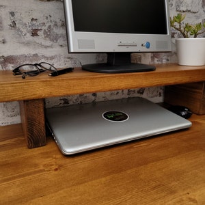 May include: A rectangular wooden monitor stand with a medium brown finish and two sturdy block legs. The stand supports a computer monitor and a small potted plant, while a silver laptop is stored in the open space underneath the shelf.