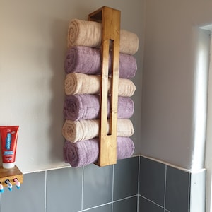 May include: A vertical wooden towel rack mounted on a wall, holding six rolled towels in alternating beige and purple colors. The rack features a simple rectangular frame design with a central vertical slat, providing storage for bathroom linens.