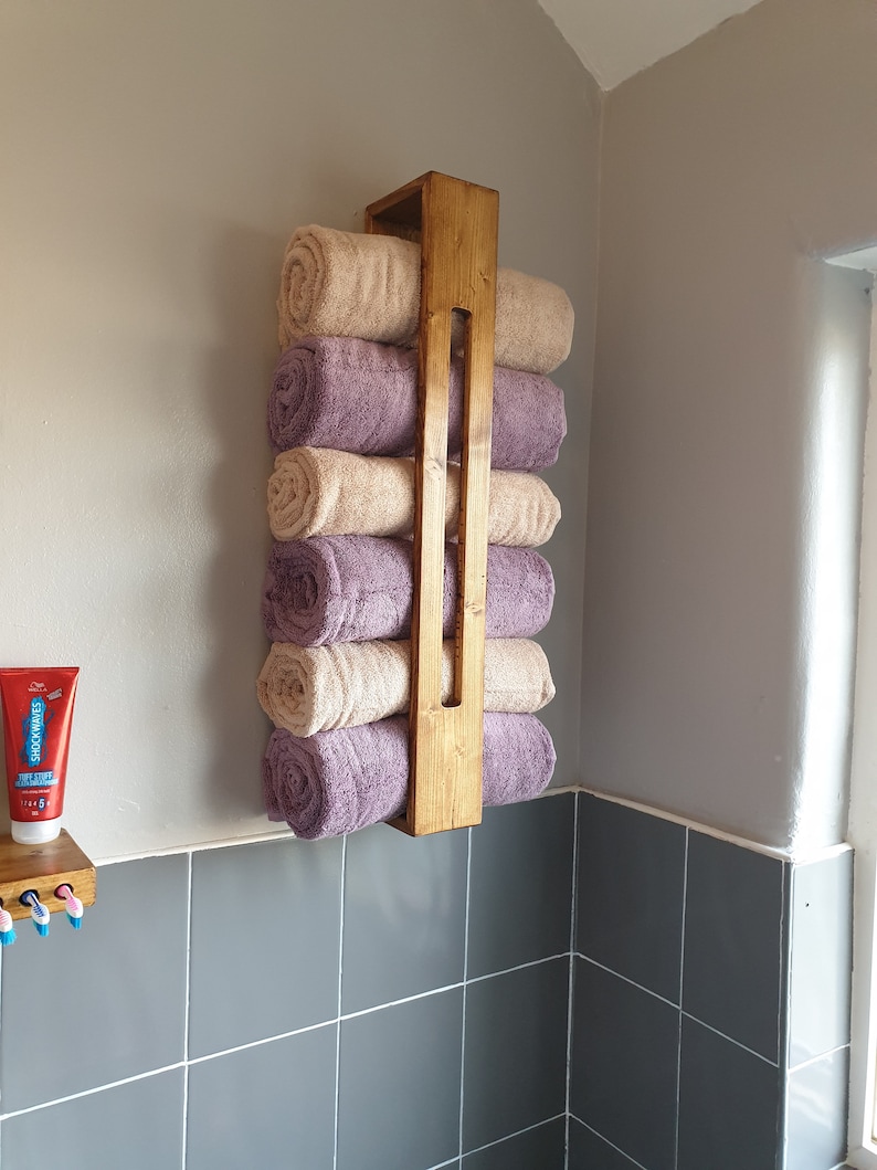 May include: A vertical wooden towel rack mounted on a wall, holding six rolled towels in alternating beige and purple colors. The rack features a simple rectangular frame design with a central vertical slat, providing storage for bathroom linens.