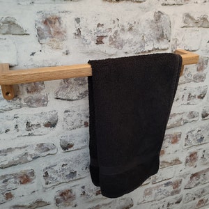 May include: A wooden towel rail with a dark grey towel hanging from it. The rail is made of light-coloured wood and is mounted on a white brick wall. The towel is folded and hangs down.