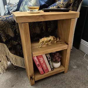 May include: A wooden side table with two shelves. The top shelf has a glass with a drink and a remote control. The bottom shelf has books and a small potted plant. A gold metal animal figurine sits on the top shelf.