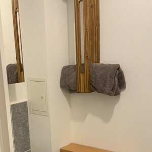 May include: A vertical rectangular wooden towel rack with a medium brown finish mounted on a wall, holding a folded gray towel. Below it sits a small gray cabinet with a light wood top, featuring open shelving and a roll of white toilet paper.