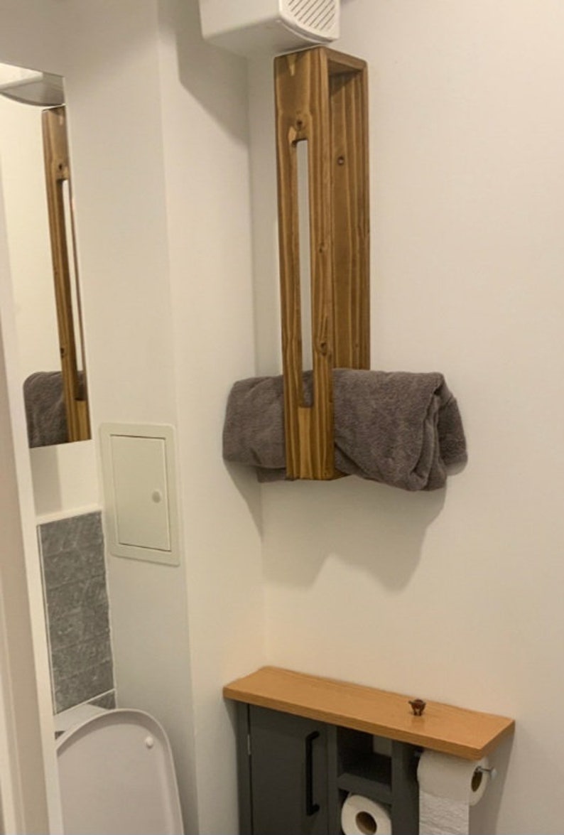 May include: A vertical rectangular wooden towel rack with a medium brown finish mounted on a wall, holding a folded gray towel. Below it sits a small gray cabinet with a light wood top, featuring open shelving and a roll of white toilet paper.