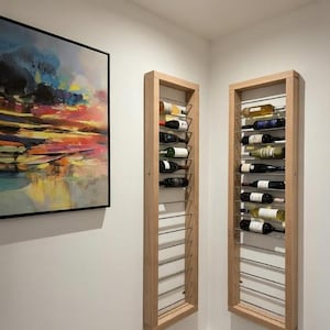May include: Two vertical, rectangular light wood wine racks mounted on white walls. Each rack features horizontal metal rods for bottle storage. The left rack holds five wine bottles, and the right rack holds seven wine bottles of various colours.