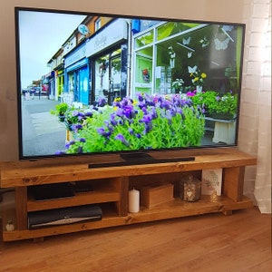 May include: A rectangular wooden television stand with a natural grain finish, featuring two open shelves. The lower shelf holds a black electronic device, a white pillar candle, a wooden box, a decorative glass container, and a small framed print.
