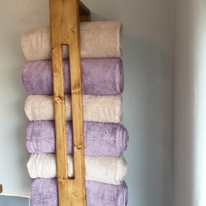 May include: A vertical wooden towel rack with a medium brown stained finish, featuring a central support beam. Six rolled towels are stacked within the frame, alternating between solid light beige and solid lavender purple colors.