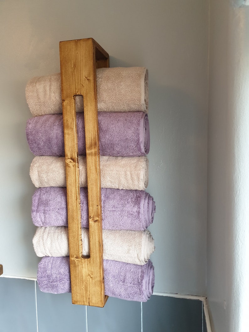 May include: A vertical wooden towel rack with a medium brown stained finish, featuring a central support beam. Six rolled towels are stacked within the frame, alternating between solid light beige and solid lavender purple colors.