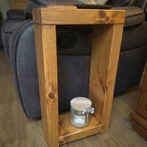 May include: A rectangular wooden side table with a medium brown stained finish and visible wood grain. The table features a flat top surface and a lower shelf holding a glass jar with a light-coloured wooden lid and a small blue ribbon detail.