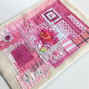 Op de afbeelding: Een handgemaakt textielkunstwerk met een patchwork-ontwerp in verschillende roze tinten. De compositie omvat vierkanten en rechthoeken van stof, versierd met knopen en decoratieve stiksels. Een bloemenprint is zichtbaar in het midden.