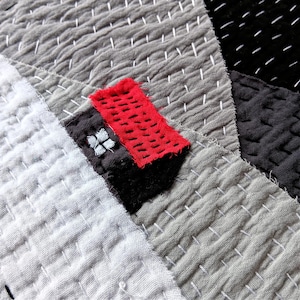 May include: Close-up of a grey, white and black fabric quilt with red and white stitching. The quilt is made of different pieces of fabric sewn together. The red fabric has a small white flower design.