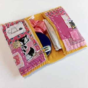 May include: Handmade fabric pouch with a yellow base and pink patchwork details. The pouch features a cat illustration, a black and white dog illustration, and a blue compact mirror. It contains a pink scrunchie, cotton buds, and a hair clip.