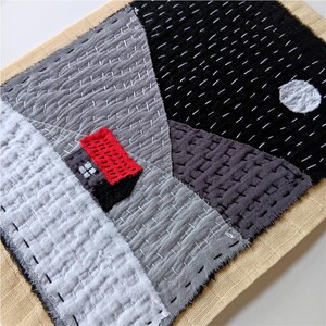 May include: A hand-stitched textile art piece featuring geometric shapes in shades of grey, black, and white. A small red rectangle is stitched in the centre of the piece, representing a house.