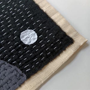 May include: A black fabric wall hanging with a white circle in the centre. The fabric is stitched with white thread in a grid pattern. The edges of the fabric are frayed and the hanging is backed with a beige fabric.