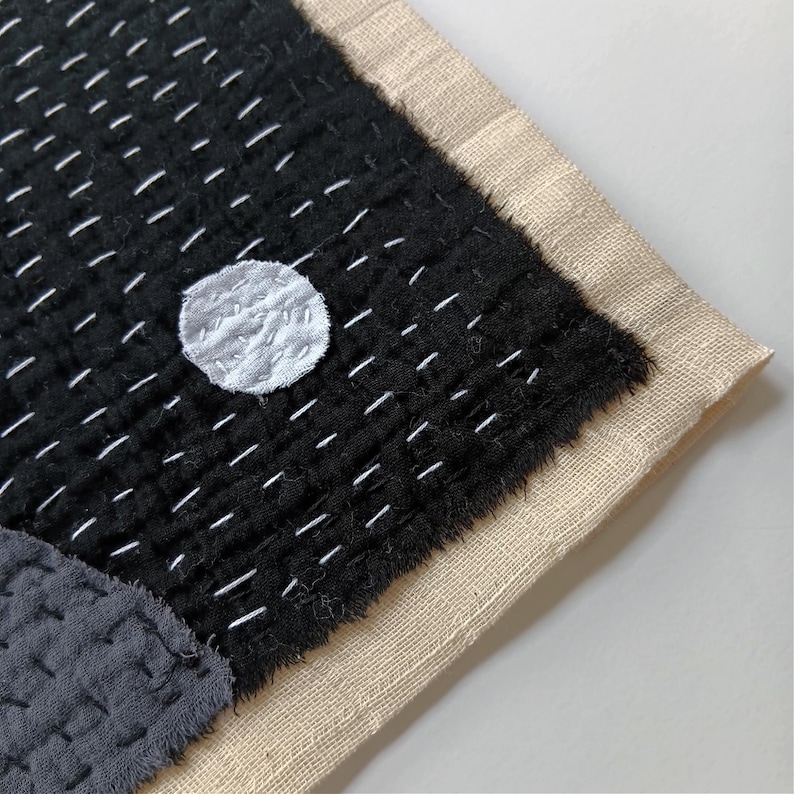May include: A black fabric wall hanging with a white circle in the centre. The fabric is stitched with white thread in a grid pattern. The edges of the fabric are frayed and the hanging is backed with a beige fabric.