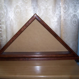 May include: A wooden triangular shadow box with a brown stain finish. The box has a shelf and is designed to display a folded American flag.