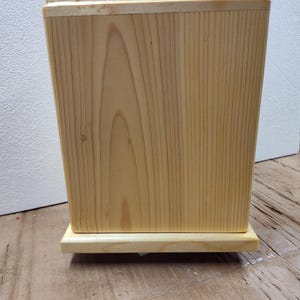 May include: A light-colored wooden box with a rectangular shape and a slightly tapered design. The box is made of wood with visible grain patterns and sits on a matching wooden base. The box has a simple, minimalist aesthetic.