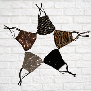 May include: Five different face masks made of sparkly fabric. The masks are in a star shape with the points facing outward. The masks are in the colors black, brown, gold, silver, and leopard print.