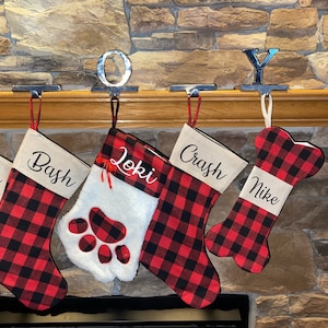 Op de afbeelding: Vier gepersonaliseerde kerstsokken van rood en zwart buffalo plaid stof. De sokken hangen aan een schoorsteenmantel met metalen haken. De sokken zijn gepersonaliseerd met de namen "Bash", "Loki", "Crash" en "Mike".