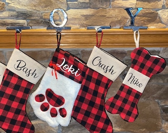 Op de afbeelding: Vier gepersonaliseerde kerstsokken van rood en zwart buffalo plaid stof. De sokken hangen aan een schoorsteenmantel met metalen haken. De sokken zijn gepersonaliseerd met de namen "Bash", "Loki", "Crash" en "Mike".