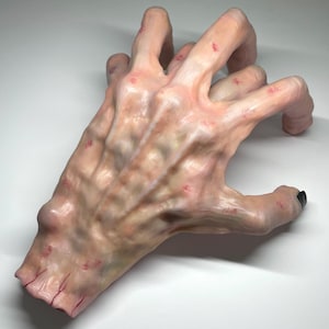 May include: A sculpted hand with a pale, textured appearance, showing visible veins and red spots. The fingers are curved, and the fingernails are painted black. The hand is set against a white background.
