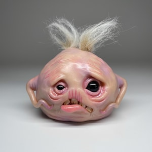 May include: A round, pale pink creature figurine with a wrinkled, fleshy texture, two small arms, and a tuft of wispy white hair on top. It features one large black eye, one smaller eye, a crooked mouth with visible teeth, and a protruding tongue.