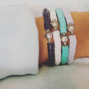 May include: Four bracelets stacked on a wrist. The bracelets are made of colorful round beads and have silver charms in the shape of seashells.