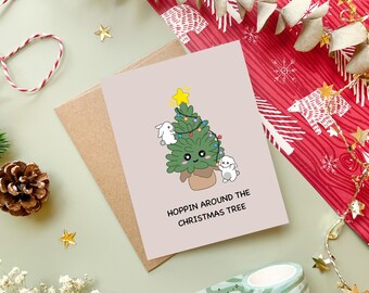 May include: A Christmas card with a cartoon Christmas tree and two white bunnies. The tree has a yellow star on top and is decorated with colorful lights. The card reads "HOPPIN AROUND THE CHRISTMAS TREE". The card is on a brown envelope.